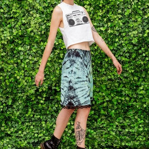 May include: A woman wearing a white crop top with a black and white boombox graphic, a tie-dye skirt, and black combat boots. She is standing in front of a green wall.