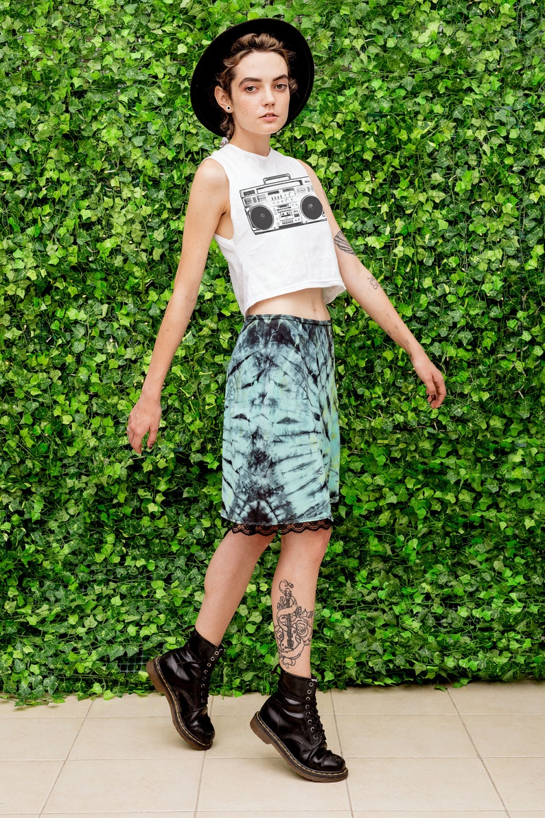 May include: A woman wearing a white crop top with a black and white boombox graphic, a tie-dye skirt, and black combat boots. She is standing in front of a green wall.