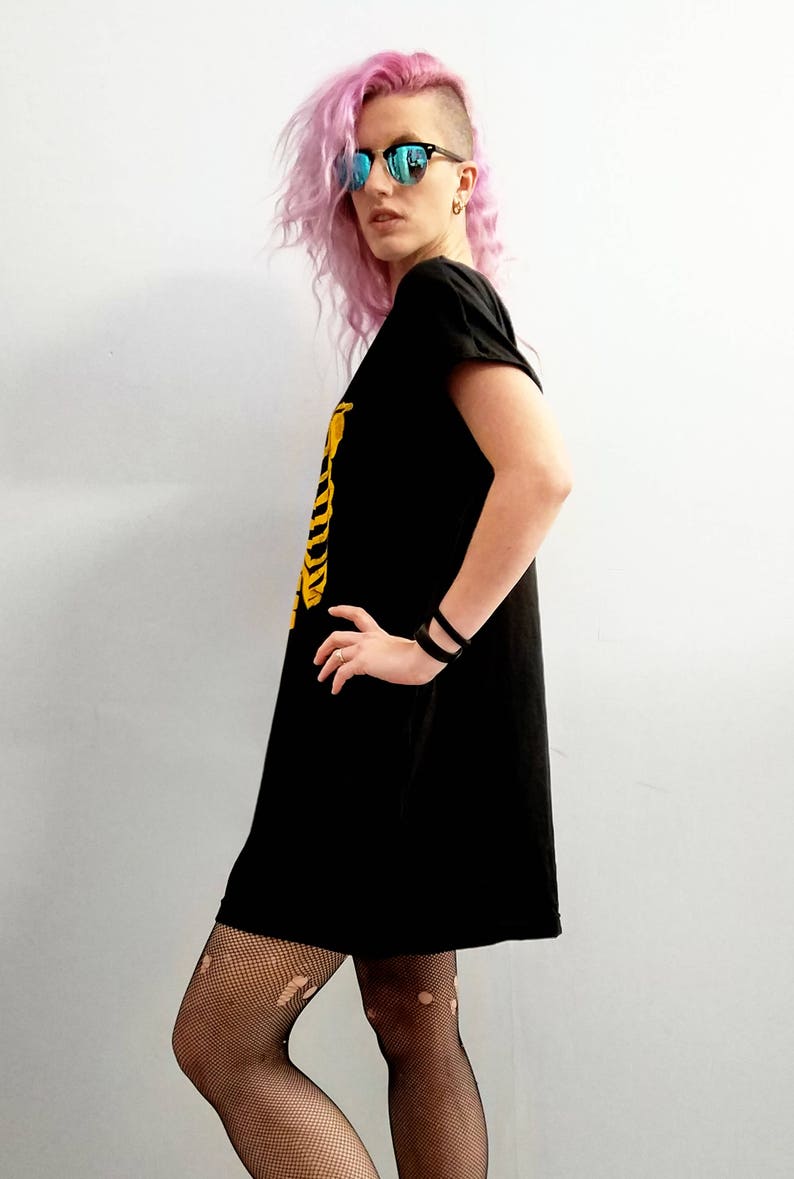 May include: A woman with pink hair and blue sunglasses wears a black t-shirt dress with a yellow skeleton graphic. She is wearing black fishnet tights.
