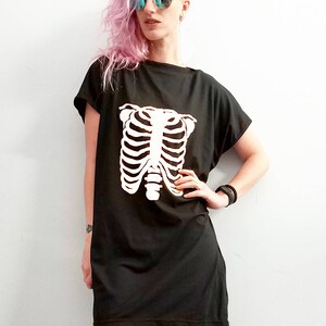 May include: A black t-shirt dress with a white skeleton ribcage graphic. The model is wearing black fishnet tights and has pink hair.