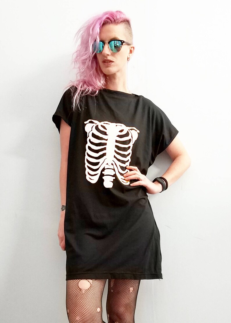 May include: A black t-shirt dress with a white skeleton ribcage graphic. The model is wearing black fishnet tights and has pink hair.