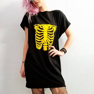 May include: A black oversized t-shirt with a yellow skeleton graphic. The model is wearing black fishnet tights and pink hair.