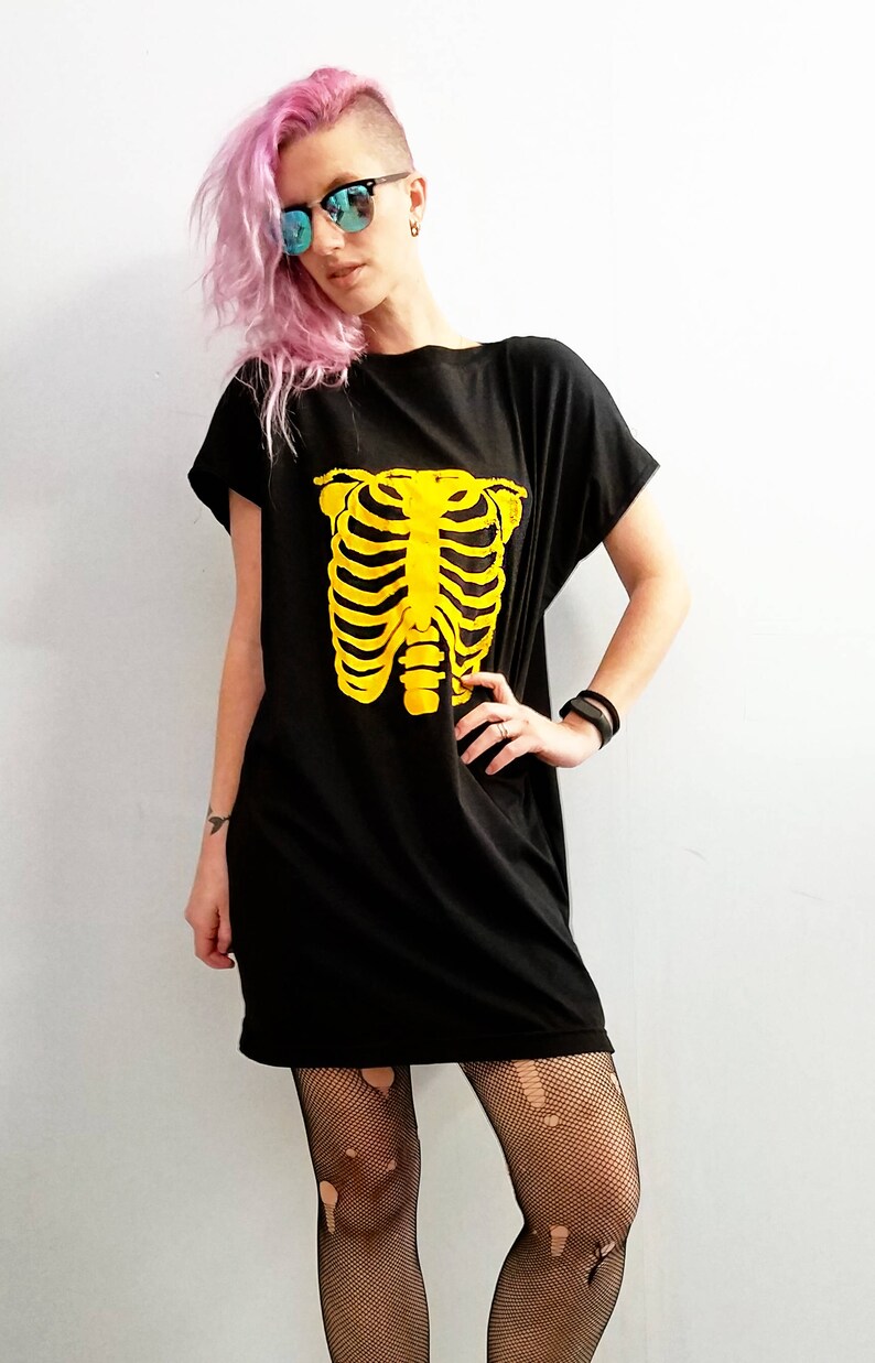 May include: A black oversized t-shirt with a yellow skeleton graphic. The model is wearing black fishnet tights and pink hair.