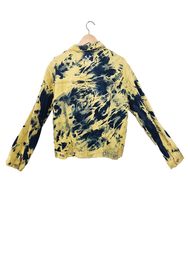 Pu&ograve; includere: Giacca di jeans gialla con motivo tie-dye blu navy. La giacca ha un colletto classico, bottoni e maniche lunghe. Un disegno ricamato bianco &egrave; sulla parte superiore della schiena. La giacca &egrave; esposta su una gruccia di legno.