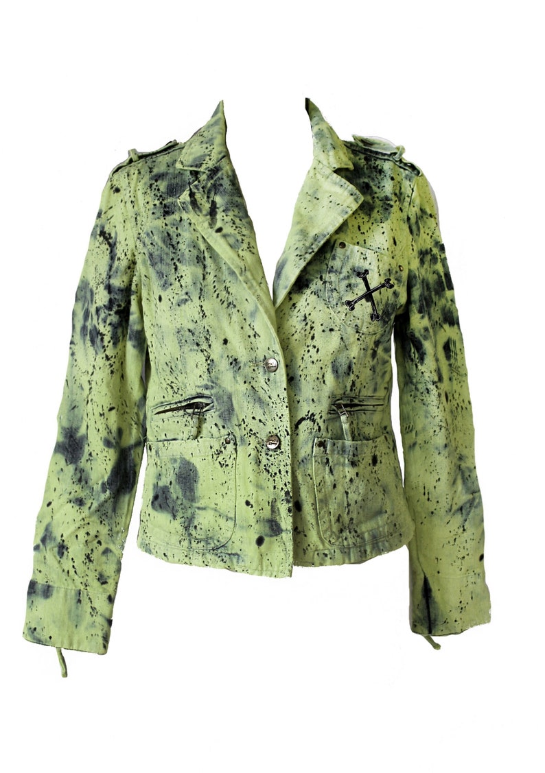 May include: A green denim jacket with black splatter paint and a silver cross detail. The jacket has a button closure and two pockets.