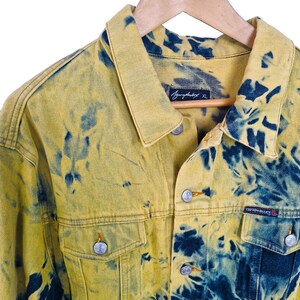Pu&ograve; includere: Giacca di jeans gialla con motivo tie-dye blu. La giacca ha una chiusura con bottoni, due tasche sul petto e un colletto. L'etichetta recita "Agurphobix XL" e "Cotton Blues". &Egrave; appesa a una gruccia di legno.