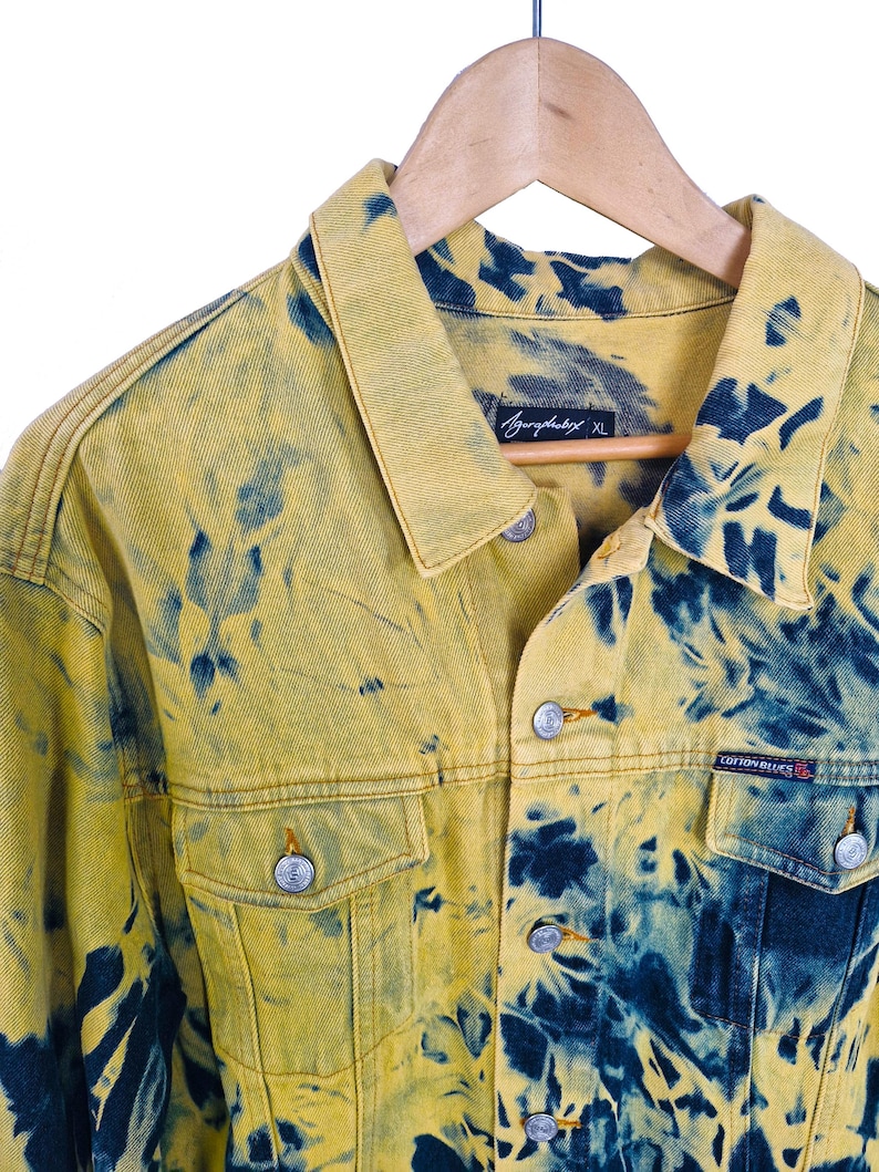 Pu&ograve; includere: Giacca di jeans gialla con motivo tie-dye blu. La giacca ha una chiusura con bottoni, due tasche sul petto e un colletto. L'etichetta recita "Agurphobix XL" e "Cotton Blues". &Egrave; appesa a una gruccia di legno.