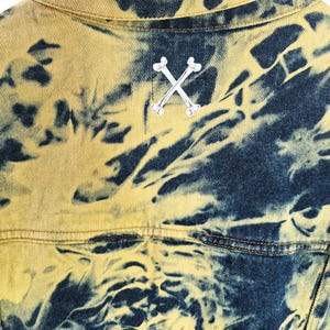 Pu&ograve; includere: Primo piano di una giacca di jeans con un motivo tie-dye unico in giallo e blu navy. La parte posteriore della giacca presenta un'emblema di ossa incrociate argentate. La giacca &egrave; appesa a una gruccia di legno.