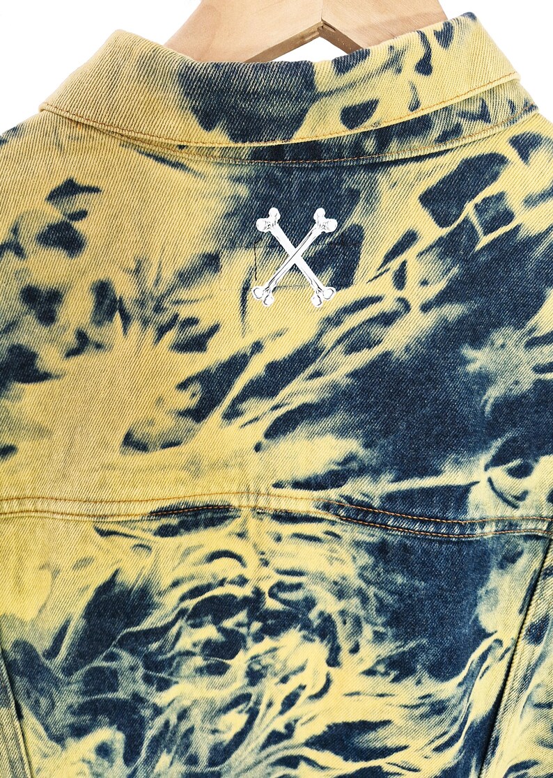 Pu&ograve; includere: Primo piano di una giacca di jeans con un motivo tie-dye unico in giallo e blu navy. La parte posteriore della giacca presenta un'emblema di ossa incrociate argentate. La giacca &egrave; appesa a una gruccia di legno.