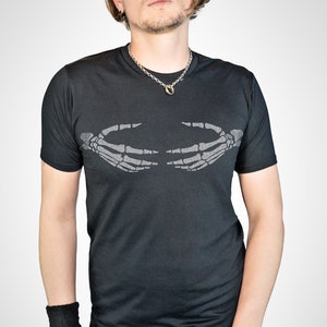 May include: A black t-shirt with a silver skeleton hand graphic on the front. The hands are facing each other and appear to be holding something. The person wearing the shirt is also wearing a silver chain necklace and a black wristband.