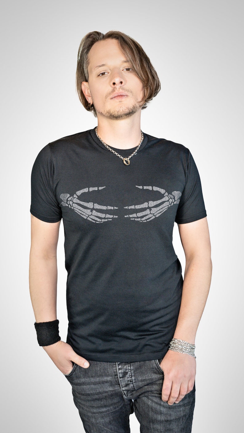 May include: A black t-shirt with a silver skeleton hand graphic on the front. The hands are facing each other and appear to be holding something. The person wearing the shirt is also wearing a silver chain necklace and a black wristband.
