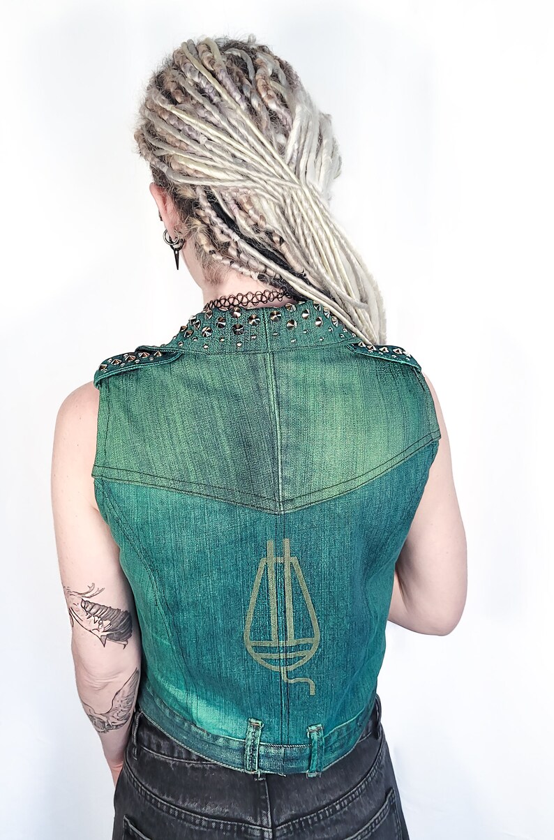 May include: A green denim vest with a gold-colored musical note design on the back. The vest has a studded collar and is sleeveless.