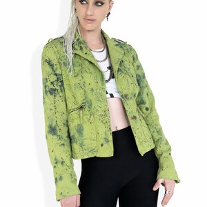 May include: A green denim jacket with black paint splatters. The jacket has a zipper closure and a button-up front. The model is wearing black pants and has dreadlocks.