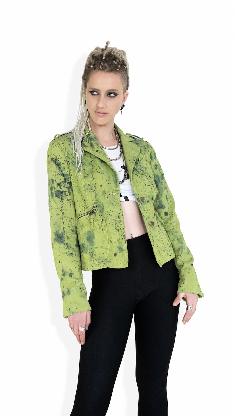 May include: A green denim jacket with black paint splatters. The jacket has a zipper closure and a button-up front. The model is wearing black pants and has dreadlocks.