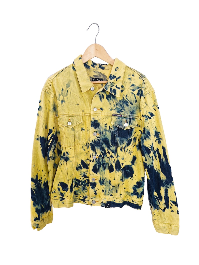 Pu&ograve; includere: Giacca di jeans gialla con motivo tie-dye blu navy. La giacca ha un design classico con chiusura a bottoni, due tasche sul petto e un colletto. &Egrave; appesa a una gruccia di legno.