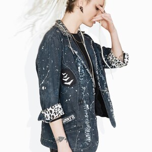 以下が含まれることがあります： 長い金色のドレッドヘアの女性は、白い塗料の飛沫と黒と白のヒョウ柄の裏地が付いた濃い青色のデニムジャケットを着ています。ジャケットの左側に3匹の白いコウモリが描かれたパッチがあり、右側に「SINKER」と書かれています。彼女は黒いジーンズを履いており、手首にタトゥーがあります。