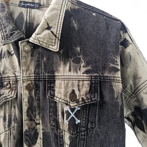 May include: A black and gray denim jacket with a unique tie-dye pattern. The jacket features a button-down front, two chest pockets, and a small embroidered design of crossed bones. The brand name "Azyrophobix" is visible on the label.