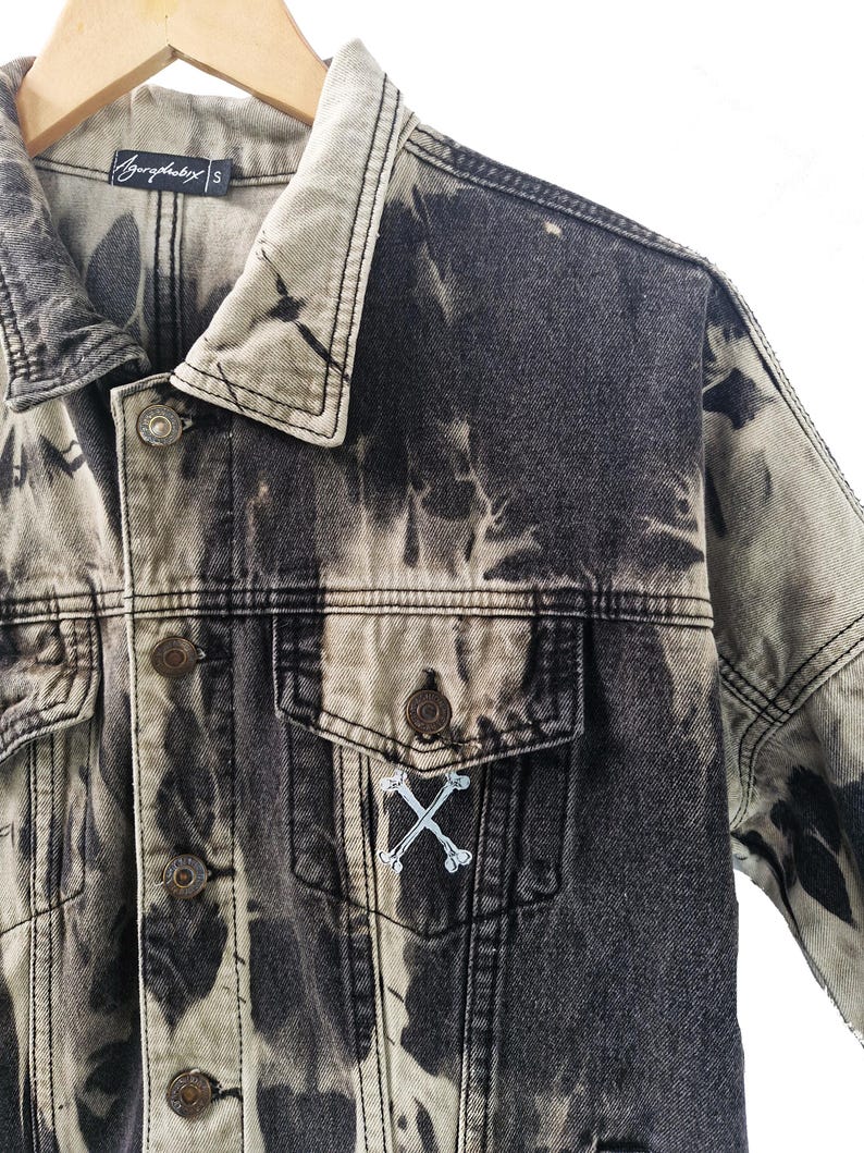 May include: A black and gray denim jacket with a unique tie-dye pattern. The jacket features a button-down front, two chest pockets, and a small embroidered design of crossed bones. The brand name "Azyrophobix" is visible on the label.