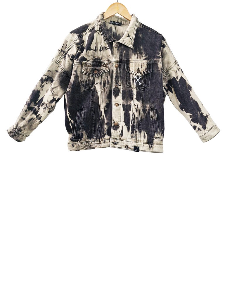 May include: A denim jacket with a unique black and white tie-dye pattern. The jacket features a classic button-up design with two chest pockets and two side pockets. A small white "X" is stitched on the front. The jacket is hanging on a wooden hanger.