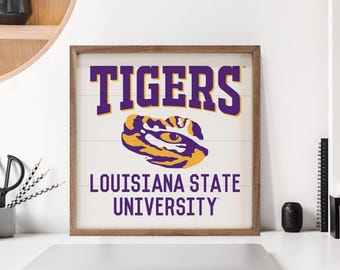 May include: Wooden framed sign with the text "TIGERS" in purple and gold, with the Louisiana State University logo. The sign is on a white background with a wooden frame. The sign is displayed on a white surface.