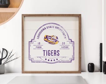 May include: Wooden framed sign featuring the Louisiana State University logo in purple and gold. The sign includes the text "Louisiana State University" and "Tigers". The sign is on a white background.
