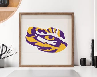 May include: Wooden framed wall art featuring a stylized eye design in purple and gold. The artwork is set against a white background with horizontal wood paneling. The frame is a natural wood color.