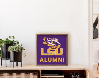 May include: A framed LSU Alumni sign featuring the university's tiger logo in gold and white on a purple background. The sign is displayed on a wooden cabinet with decorative plants.