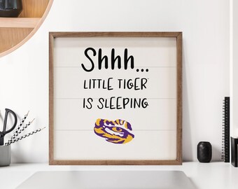 May include: Wooden framed sign with the text "Shhh... LITTLE TIGER IS SLEEPING" in black lettering. The sign features a purple and gold tiger graphic. The background is white with horizontal wood grain lines.