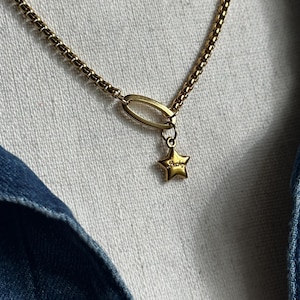 May include: A gold-toned necklace featuring a box chain and a central oval link. A small, five-pointed star charm hangs from the oval link. The star charm has the word "Lucky" engraved on its surface in a cursive script, adding detail to the piece.