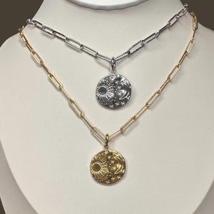 May include: Two necklaces with celestial pendants. One necklace is gold-toned with a sun, moon, and star pendant. The other necklace is silver-toned with a similar pendant. Both necklaces have a chain link design.