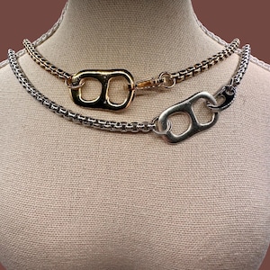 Puede incluir: Dos collares con un diseño único. Uno es dorado, el otro plateado. Cada uno presenta una cadena y una gran anilla de lata estilizada como punto focal. Los collares se exhiben en un maniquí.