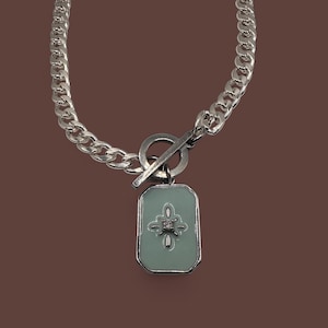 Puede incluir: Collar de cadena plateada con un colgante rectangular. El colgante tiene un fondo verde claro con un diseño floral plateado y una pequeña gema brillante en el centro. El collar presenta un cierre de palanca.