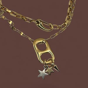 Puede incluir: Un collar dorado de dos capas. La capa superior tiene una cadena con eslabones rectangulares y un cierre de imperdible. La capa inferior presenta un colgante de anillo grande con dijes de estrella y punta. El fondo es marrón liso.