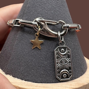 May include: Silver-toned bracelet with a large clasp and two charms. One charm is a gold-colored star, and the other is a rectangular pendant with an eye, sun rays, moons, and stars design. The bracelet is displayed on a gray surface.
