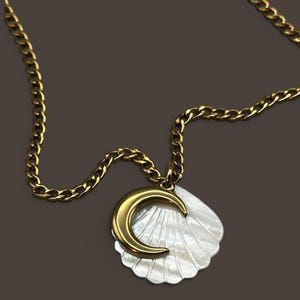 May include: A gold-toned metal chain necklace featuring a pendant composed of a white iridescent seashell layered with a smooth, curved gold-toned crescent moon shape. The necklace has a classic link chain design and a polished metallic finish.