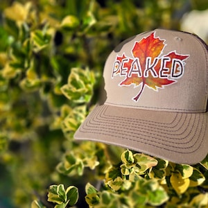 May include: A tan and brown baseball cap with the word "PEAKED" and a colorful maple leaf design. The hat has a curved brim and is set against a backdrop of green and yellow foliage.