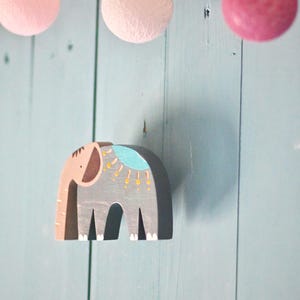 Op de afbeelding: Een houten olifantenfiguur geschilderd in tinten grijs, roze en blauw met gele decoratieve accenten. De olifant heeft een gestileerd ontwerp met een roze slurf, een grijs lichaam en een blauw zadelachtig vlak met gele stippen.
