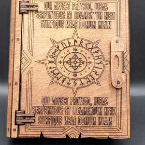 May include: Wooden box with laser-etched design. The box features a central circular emblem with runes and text in a decorative border. The text "QUI AFFECT PROTEGO, IUBAS SERPENTIBUS ET LIGAMENTUM MEIS STIRPIQUE MEAE DOMUM MEAM" is etched on the box.