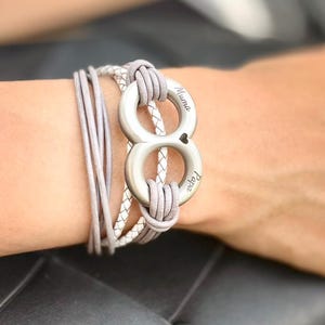 Könnte beinhalten: Ein mehrsträngiges Armband mit hellgrauen und weißen geflochtenen Kordeln, die von einem silberfarbenen, unendlichkeitsförmigen Metallanhänger zusammengehalten werden. Der Anhänger trägt die Gravur Muma und Papa mit einem Herz.