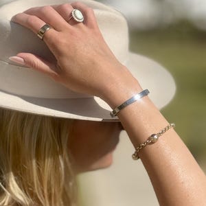 Op de afbeelding: Een zilverkleurige armband, een goudkleurige kettingarmband met een centrale kraal, een brede zilverkleurige ring en een ring met een ovale witte steen omringd door kleine heldere steentjes. De sieraden worden gedragen met een beige vilten fedora hoed.