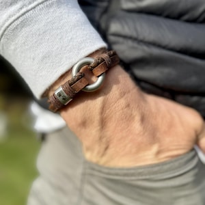 May include: A brown leather braided bracelet featuring a central matte silver-toned metal ring and a rectangular silver-toned metal bead engraved with two small paw prints. The bracelet is constructed from multiple strands of leather cord.