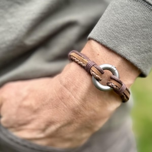 Pode incluir: Uma pulseira de couro castanho com fecho de metal prateado. A pulseira apresenta detalhes em couro trançado e costurado. O fecho é um grande anel circular. A pulseira é usada no pulso.