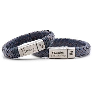 Op de afbeelding: Twee blauwe leren armbanden met zilveren metalen sluitingen. De sluitingen zijn gegraveerd met de namen "Nova" en "Frankie" en poot afdrukken. De armband met de naam "Frankie" heeft ook de woorden "With me always" gegraveerd.