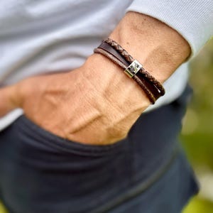 Può includere: Un braccialetto in pelle marrone con una treccia e una chiusura argentata. Il braccialetto è indossato su un polso, con la mano della persona in tasca. La chiusura ha un design a impronta di zampa.