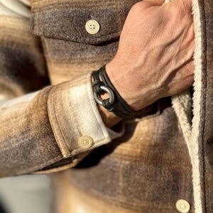 May include: A dark olive green paracord bracelet featuring a circular metallic clasp. The bracelet is constructed from multiple strands of woven cord that wrap around the wrist, secured by a smooth, matte-finished silver-toned metal ring.
