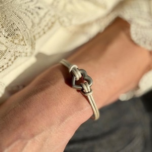 Könnte beinhalten: Ein Armband mit einer hellbeigen Kordel und einem mittigen, silberfarbenen Herzanhänger aus Metall. Die Kordel ist durch das Herz geführt und bildet ein schlichtes Design, das sich um das Handgelenk legt. Das Metallherz hat eine glatte, matte Oberfläche.