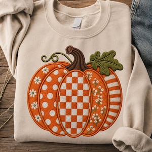 Puede incluir: Una sudadera de cuello redondo en color beige con un gran diseño de calabaza bordado en la parte delantera. La calabaza está dividida en secciones con patrones naranjas y blancos que incluyen lunares, cuadros, flores y rayas.