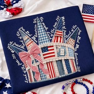 May include: A navy blue decorative pillow featuring a patriotic crown design. The crown is adorned with red, white, and blue fabric patches, a gingham bow, and lace trim. Text includes "Land of the free" and "250 years of freedom."