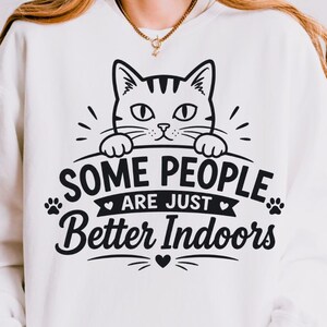 May include: A white crewneck sweatshirt featuring a black graphic of a cat peeking over a line. The text reads, "SOME PEOPLE ARE JUST Better Indoors," accompanied by small heart and paw print icons. The design is centred on the chest area.