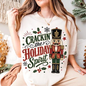 May include: A cream-colored t-shirt featuring a graphic of a nutcracker in a red, green, and black uniform. The text "CRACKIN' the HOLIDAY SPIRIT" is printed in green and red script and block lettering, surrounded by holly, berries, and snowflakes.